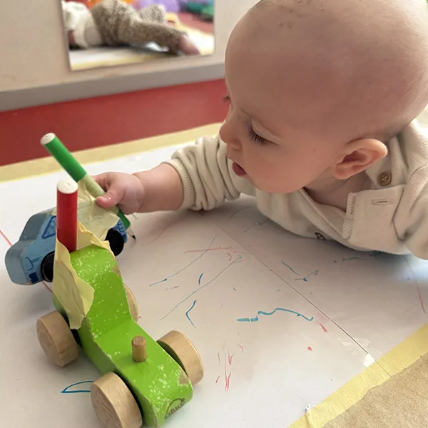 Un enfant à la crèche La Nanosphère à Lausanne dessine avec une voiture équipée d’un feutre sur une grande feuille au sol.